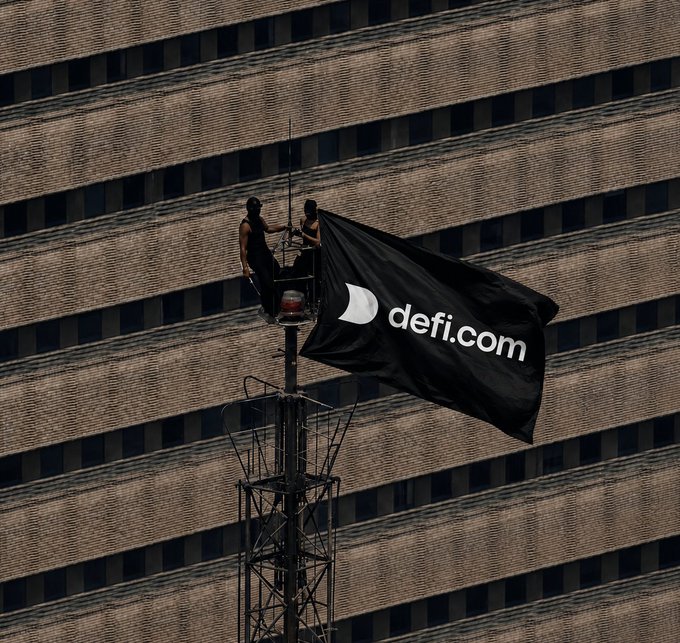 Two people sit atop a tall tower/cell mast against a brick building facade, unfurling a large black flag with the white 'defi.com' logo.