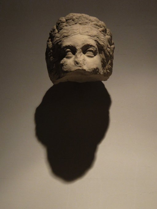 An ancient carved stone head sculpture mounted on a wall, casting a dramatic shadow under spot lighting, likely in a museum setting.