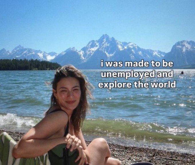 A smiling young woman sits on a pebbled beach with snow-capped mountains and a lake behind her, with overlaid text reading 'i was made to be unemployed and explore the world'.