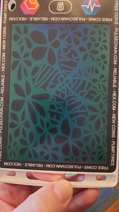 A photograph of a hand holding a PulseChain branded card with a decorative cut-out floral pattern revealing a teal backing, surrounded by text reading 'FREE COINS · PULSECHAIN.COM · RELIABLE · HEX.COM · NEW COINS · PUMPTIRES'.