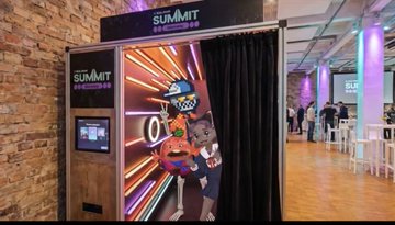 The image shows a large, colorful photo booth with a cartoon character inside, likely representing a mascot or promotional figure for an event like the Berlin Summit. The booth has a black curtain covering its interior, adding to the overall visual appeal of the scene. There are several people in the background, possibly attending the summit or participating in some sort of event at the venue.