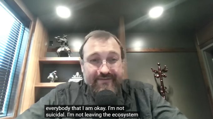 A webcam video still of a bearded man with glasses speaking into a microphone in a room with figurines on shelves, with captions reading 'everybody that I am okay. I'm not suicidal. I'm not leaving the ecosystem'.