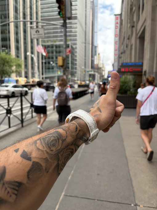 A first-person view of a tattooed arm wearing a white watch, giving a thumbs-up while standing on a New York City sidewalk near the NBC Studios and Radio City Music Hall.