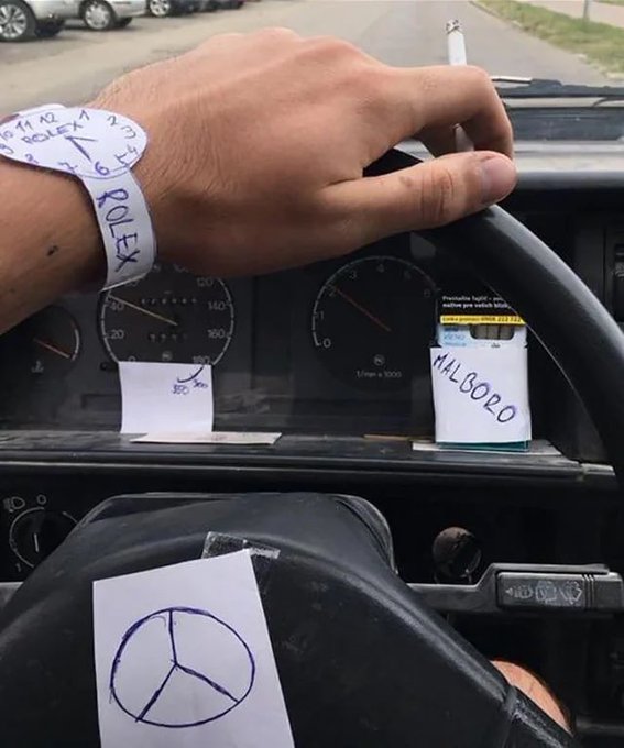 A first-person view from inside a Mercedes-Benz showing a driver's hand on the steering wheel wearing a 'ROLEX' wristband, with handwritten notes taped to the dashboard reading 'HLBOBO' and showing a Mercedes logo.