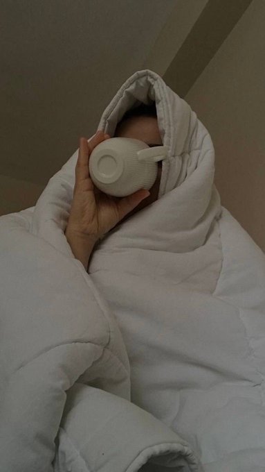 A person lies in bed wrapped snugly in white bedding, holding a white ceramic mug up to their face.