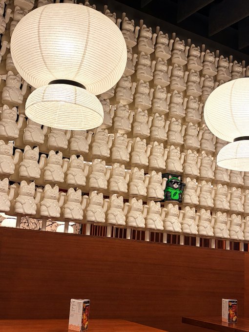 An interior space decorated with numerous small white cat figurines arranged on a wall, illuminated by paper lantern pendant lights, with one distinctive green figurine among them.