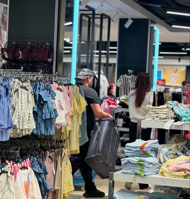 A shopper wearing a cap browses through racks of pastel-colored children's clothing in a retail store, with handbags displayed on shelves above.