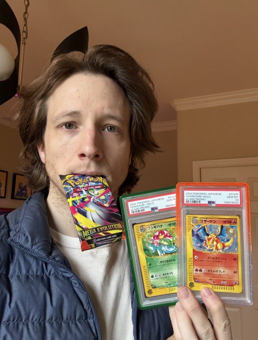 A man with shoulder-length hair wearing a navy jacket holds a Pokémon Trading Card Game Mega Evolution booster pack in his mouth and displays two PSA-graded Japanese Pokémon cards, including a Venusaur (1996) and Charizard (1999) card.