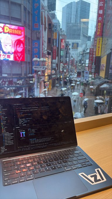 A laptop sits on a desk by a window overlooking a rainy, neon-lit city street in Japan, with code visible on the screen.