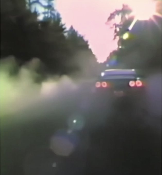 A blurry, low-light image showing the rear of a sports car with glowing red taillights on a road at dusk, with significant lens flare and motion blur.