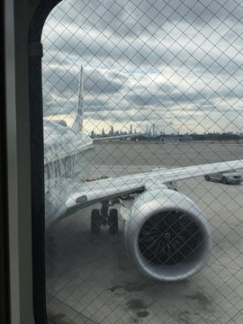 A view through a wire mesh window showing an airplane engine and wing on the tarmac, with a city skyline visible in the distance under a cloudy sky.