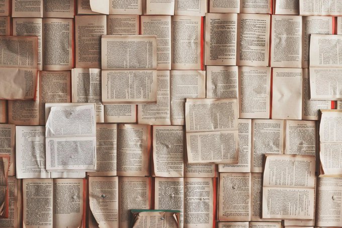 A wall covered in overlapping open book pages creates a textured mosaic of dense text, with no legible words or numbers visible at this resolution. The image's overwhelming display of printed pages visually amplifies the post's critique of excess content generation, illustrating the "madness" of endless text production that a slowdown in tokenmaxxing would supposedly curb.
