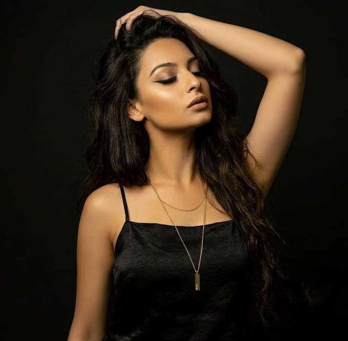 A professional studio portrait of a woman with long dark hair posing with one arm raised behind her head, wearing a black camisole and layered gold necklaces against a black background.