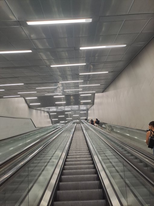 A photograph looking down a long, empty escalator descending into an underground space, illuminated by parallel rows of bright rectangular ceiling lights, with a couple of people visible in the distance.