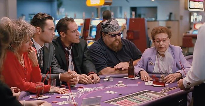 A diverse cast including an elderly woman in lavender, two men in suits, and a man with a bandana sits at a casino card table, surrounded by chips, drinks, and a dealer, evoking the unpredictable gamble highlighted in the post's commentary on coin investing.