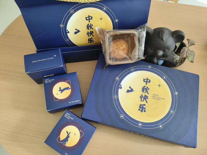 A Mid-Autumn Festival gift set featuring a large yellow moon design and '中秋快乐' (Happy Mid-Autumn) text, accompanied by a mooncake, small navy blue boxes, and a black mouse-shaped container.