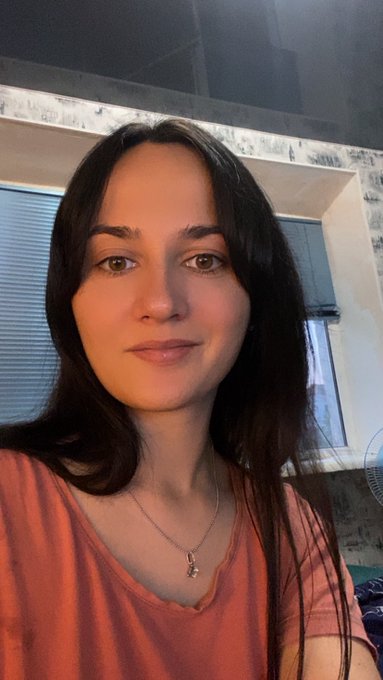 A selfie of a woman with long dark hair wearing an orange t-shirt and a small pendant necklace, taken indoors near a window with blinds.