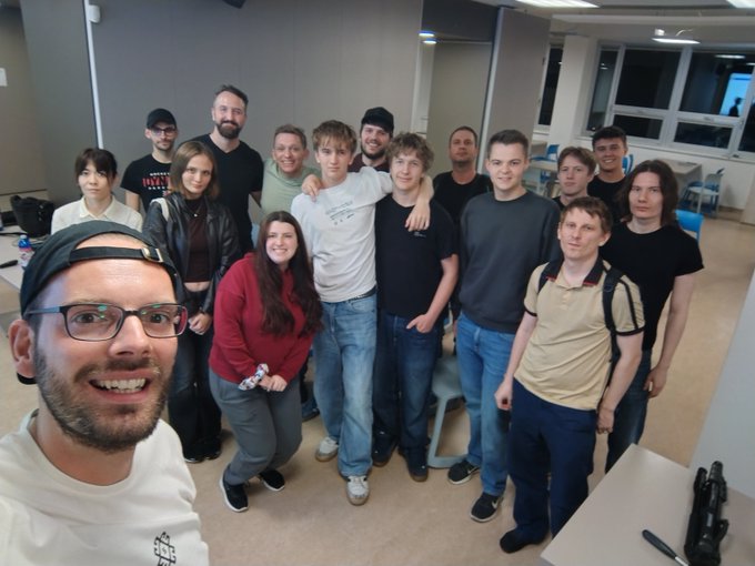 A selfie of a smiling man in a white shirt and cap with a group of approximately 14 others posing together indoors.