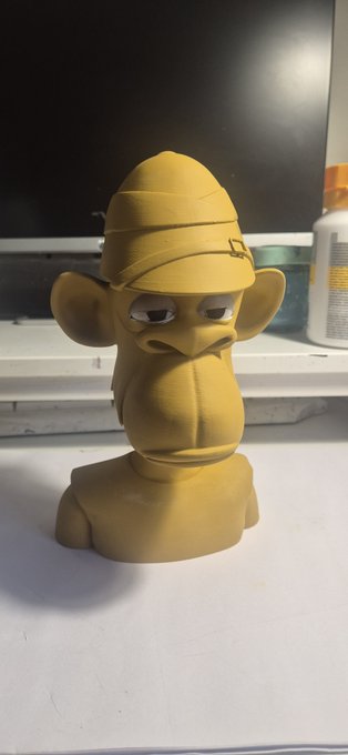 A handmade yellow clay sculpture of a grumpy-looking monkey character wearing a hat, sitting on a white surface in front of a monitor.