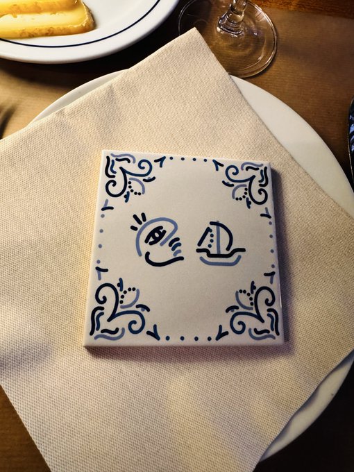The image features a white plate with a blue and white tile design, placed on top of a beige tablecloth. The plate has a small white napkin folded around it, adding an elegant touch to the setting. In the background, there is another plate holding food, suggesting that this might be part of a dinner or gathering. The image also contains text that reads "Azulejo @EfdotStudio at the @opensea @shillrxyz @LoreStudio_io dinner".