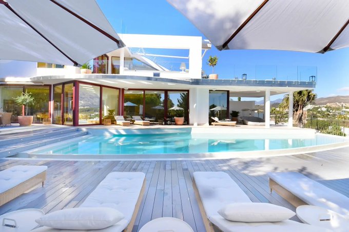 A luxurious modern white villa with a large swimming pool, sun loungers, and shaded umbrellas overlooks a scenic landscape, evoking the upscale maritime lifestyle that Ethra Harbor's onchain yield and RWA tokenization efforts aim to bring to the maritime industry.