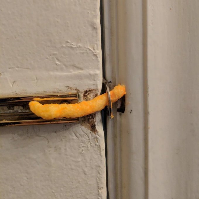 A close-up of a single orange, cheese-flavored corn puff snack threaded onto a metal keyhole escutcheon plate, locking a white door.