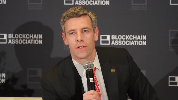 A man in a dark suit, white shirt, and red patterned tie speaks into a microphone at a Blockchain Association event, with the organization's logo visible on the backdrop behind him.