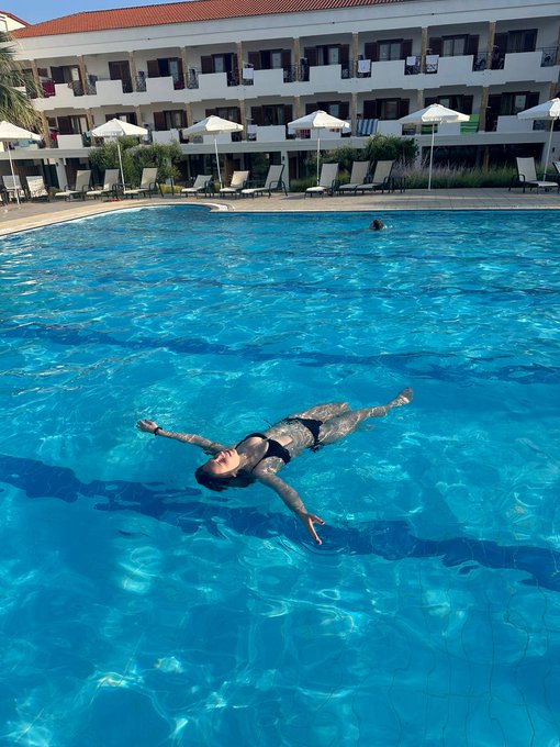 A person with tattoos floating on their back in a clear blue swimming pool at a resort, with a hotel building, sun loungers, and umbrellas visible in the background.