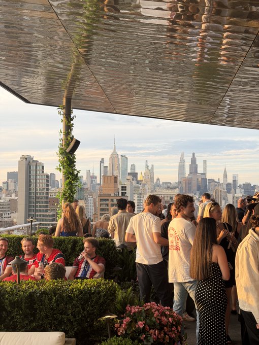 The image shows a group of people standing outside, possibly at an event or gathering. They are positioned under a large metal structure that provides shade for them. The scene takes place near a city skyline with tall buildings visible in the background. There is also a potted plant and some flowers nearby, adding to the outdoor atmosphere. The image does not provide any information about the cost of the event or the people's attire, but it seems like an informal gathering where everyone appears to be enjoying themselves.