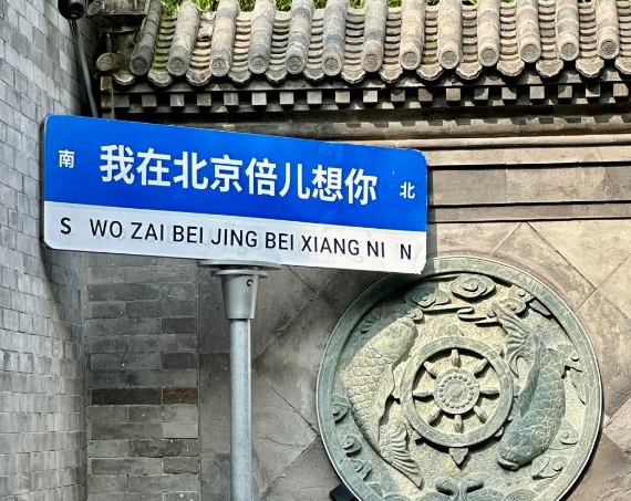 A blue and white street sign in Beijing with Chinese characters '我在北京倍儿想你' and pinyin 'WO ZAI BEI JING BEI XIANG NI', marked with 南 (S) and 北 (N) directional indicators, mounted in front of traditional gray brick architecture and a carved stone relief.