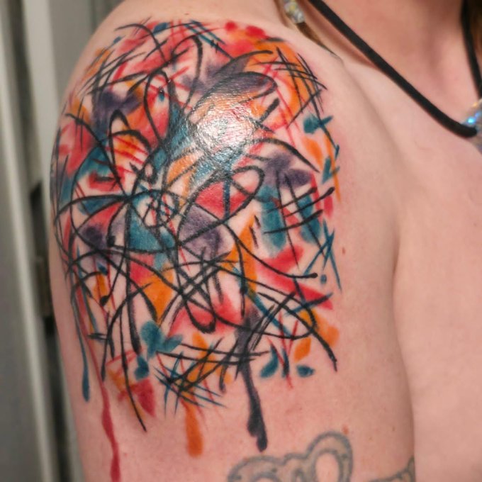 A colorful watercolor-style tattoo on a person's upper arm featuring abstract splashes of red, orange, blue, and purple with black scratchy line work over the top.