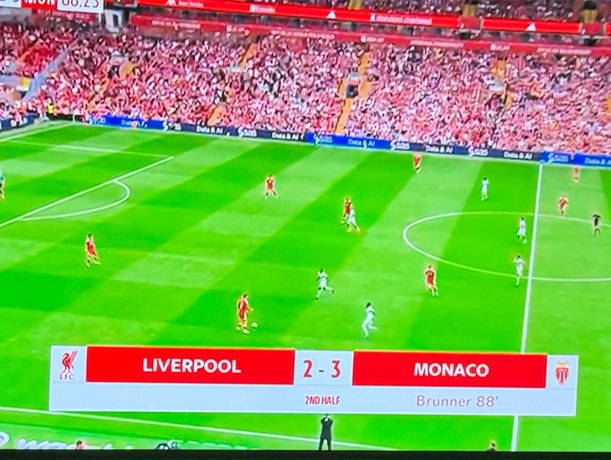 A football match is underway at Anfield, with the scoreboard showing Liverpool trailing Monaco 2-3 in the second half, with Monaco's Brunner having scored in the 88th minute, while players from both teams are positioned across the green pitch in front of a packed stadium crowd.