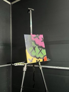 An abstract painting on a canvas mounted on a metal easel against a dark grey wall, featuring bright pink, green, and yellow colors.