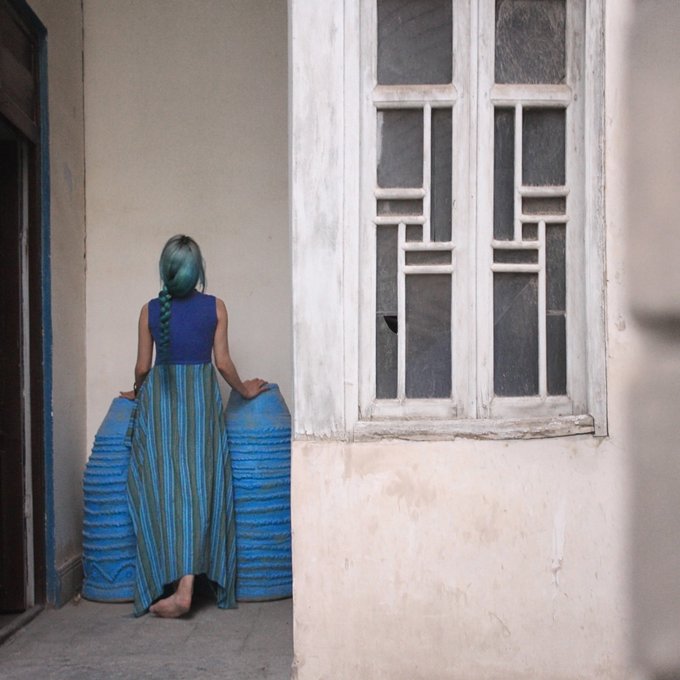 A photograph of a barefoot woman with a long blue braid, wearing a blue and green striped dress, standing with her back to the camera next to a whitewashed window.