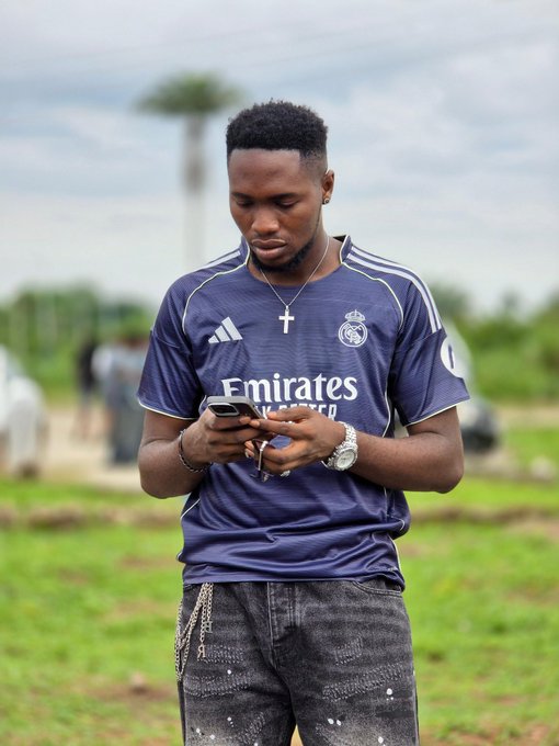 A photograph shows a young man wearing a navy Adidas Real Madrid 'Emirates Fly Better' jersey, looking down at his phone while standing in a grassy field.