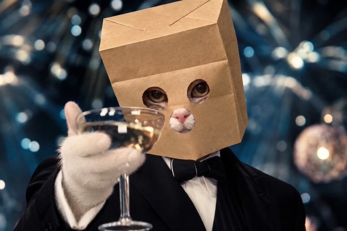 A humorous image of a cat wearing a brown paper bag on its head with eye holes cut out, dressed in a black tuxedo and holding a glass of wine, set against a blue bokeh background.