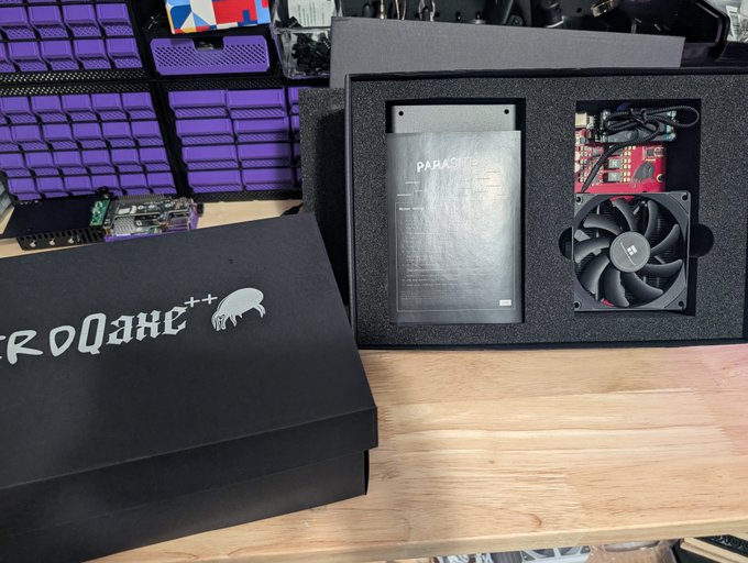 A black box labeled "NerdQAxe++" sits open on a wooden desk, revealing a Parasite-branded mining ASIC and a cooling fan nestled in protective foam, alongside what appears to be a control board. The image showcases the newly arrived Bitcoin mining hardware being unboxed, ready to be added to the miner's fleet through the parasite.space platform.