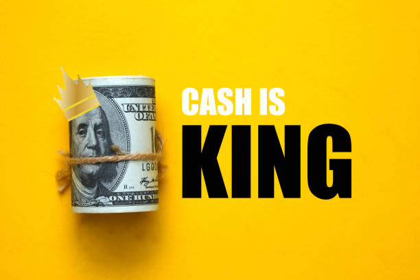 A rolled-up US one hundred dollar bill adorned with a paper crown and twine sits against a yellow background next to bold black text reading "CASH IS KING".