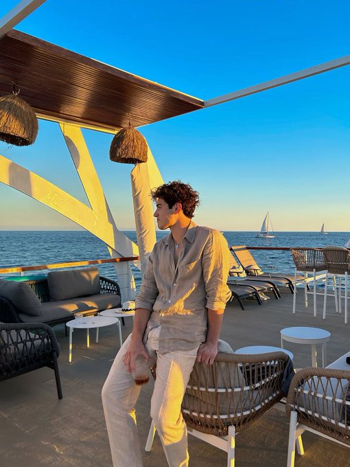 A young man in a beige linen outfit leans against a wicker chair at an oceanfront lounge during golden hour, with sailboats visible in the background.