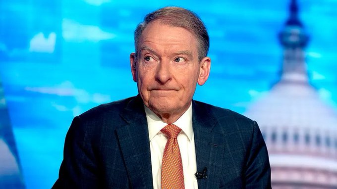A man in a dark suit with an orange tie appears to be on a news show with a blue background featuring an image of the US Capitol building.