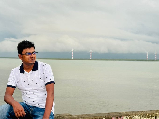 The image features a man sitting on a wall by the water with his back to the camera. He's wearing a white polo shirt, blue jeans, and glasses. The scene takes place near a body of water where several wind turbines can be seen in the distance. The man appears to be lost in thought or daydreaming while enjoying the view.