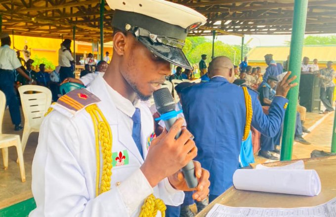 A man in a white military or cadet uniform with gold cord accents and a peaked cap speaking into a microphone at what appears to be an outdoor event with graduates in blue gowns in the background.