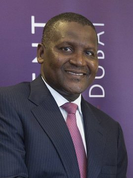 A man in a dark suit and pink tie smiles in front of a purple background with 'DUBAI' visible in white text.