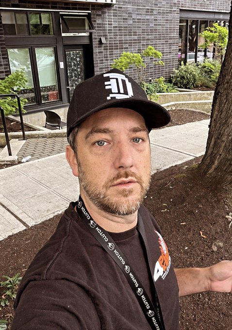 A man wearing a black cap and a lanyard that repeatedly reads 'JUJUTSU' takes a selfie outside a building.