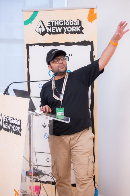The image features a man standing behind a podium, giving a presentation or speech at an event. He has his hand raised, possibly emphasizing a point he's making. The background of the image shows a banner with the text "ethGlobal New York" written on it and another banner that says "Get Started with Canton Network, Writing Daml, and Deploying on DevNet". This suggests that the man is speaking at an event related to technology or business, possibly discussing topics such as software development, networking, or project management.