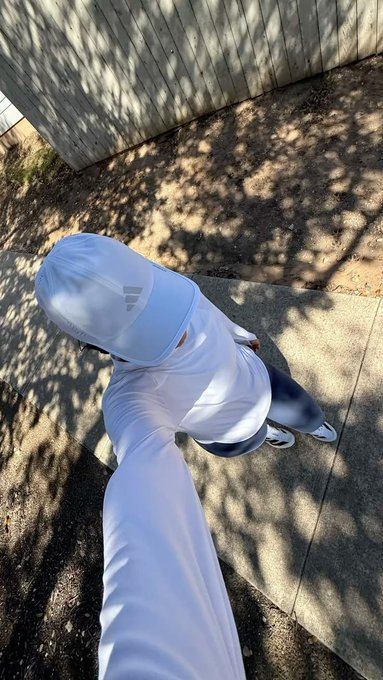 A high-angle view of a person wearing a white hooded shirt and grey pants standing on a paved path in a shaded outdoor area.