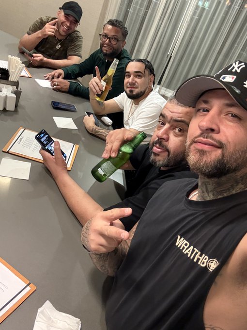 Five men pose together at a restaurant table, with one wearing a "WRATHBO" tank top and a New York Yankees cap, while others hold a Corona bottle and a Dos Equis—capturing a candid family-style gathering among close friends.
