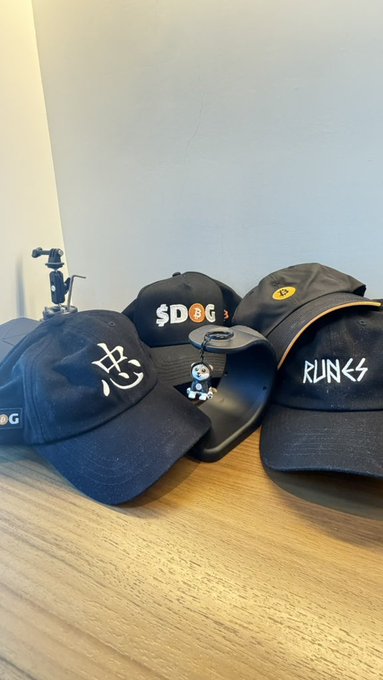 A collection of black baseball caps sits on a wooden surface, featuring logos and text including "SDOG", "忠", and "RUNGS".