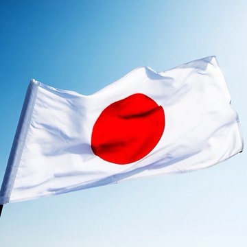 The image shows the Japanese flag—a white banner with a bold red circle—billowing against a clear blue sky, symbolizing Japan in the context of news about the JPYC stablecoin pilot at Lawson convenience stores. The flag visually anchors the announcement of Japan's upcoming stablecoin payment trial.