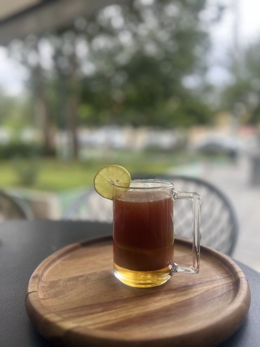 A glass mug containing a dark tea-like beverage garnished with a lime slice sits on a wooden tray on an outdoor table.